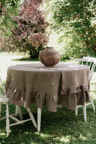 Apaļš lina galdauts ar volāniem kapučīno krāsā uz dārza galda ar diviem koka krēsliem zaļā dārzā
 Round linen tablecloth with ruffles in cappuccino color on a garden table with two wooden chairs in a green garden