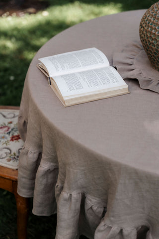 Apaļš lina galdauts kapučīno krāsā ar volāniem uz dārza galda, uz tā atvērta grāmata
Round cappuccino linen tablecloth with ruffles on a garden table with an open book on top