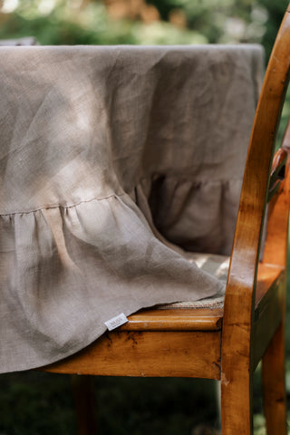 Kapučīno krāsas lina galdauts ar volāniem un NUSS etiķeti, pārkārts pār koka krēslu
Cappuccino linen tablecloth with ruffles and NUSS label draped over a wooden chair