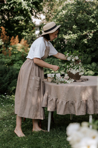 Kapučīno krāsas apaļš lina galdauts ar volāniem uz dārza galda, dekorēts ar keramikas vāzi un hortenzijām
Cappuccino round linen tablecloth with ruffles on a garden table, decorated with a ceramic vase and hydrangeas