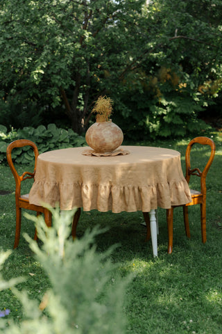 Apaļš lina galdauts karameļu krāsā ar volāniem uz galda zaļā dārzā, divi koka krēsli | Round caramel linen tablecloth with ruffles on a garden table, two wooden chairs