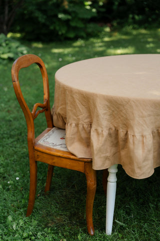Apaļš karameļu lina galdauts ar volāniem uz apaļa galda dārzā, blakus koka krēsls | Round caramel linen tablecloth with ruffles on a round garden table, wooden chair beside
