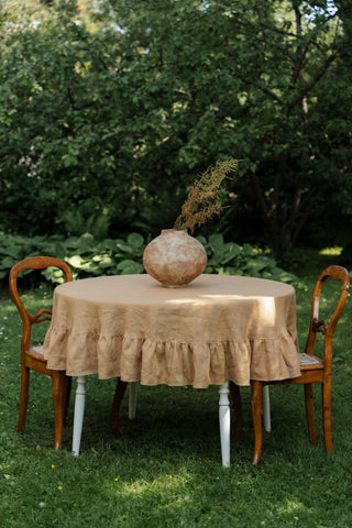 Apaļš lina galdauts karameļu krāsā ar volāniem uz apaļa galda dārzā, dekorēts ar māla vāzi un sausiem ziediem | Round caramel linen tablecloth with ruffles on a garden table, decorated with a clay vase and dried flowers