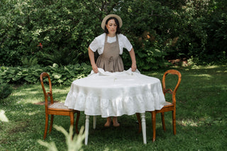 Balts lina galdauts ar volāniem un sieviete ar priekšautu klāj galdautu uz galda, dārzā / White raound linen tablecloth with ruffle in garden with antique chairs and women in retro apron