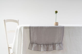 gaiši pelēks lina celiņš ar volānu uz galda ar ziedu