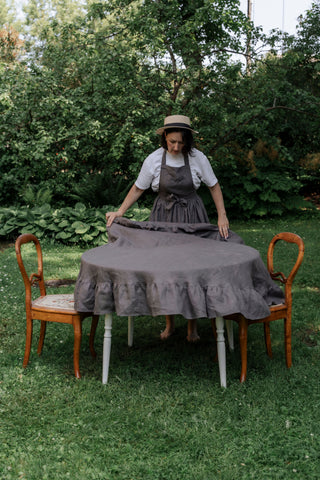 	Sieviete liek pelēku apaļu lina galdautu ar volāniem uz galda āra vidē	Woman placing grey round linen tablecloth with ruffles on outdoor table