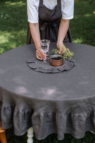 Sieviete kārto pelēku lina galdautu ar volāniem uz apaļa galda dārzā	Woman arranging grey linen tablecloth with ruffles on round garden table
