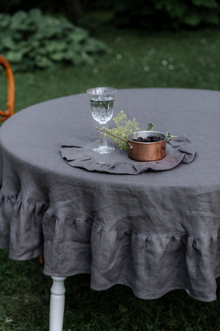 Apaļš pelēks lina galdauts ar volāniem uz balta galda dārzā ar kristāla glāzi un vara katliņu	/ Round grey linen tablecloth with ruffles on a white garden table with crystal glass and copper pot