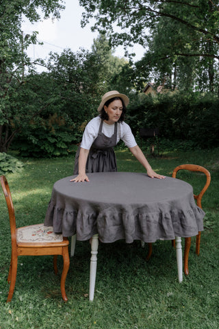 	Sieviete ar cepuri pie apaļa galda ar pelēku lina galdautu ar volāniem zaļā dārzā	Woman in hat standing by round table with grey linen ruffle tablecloth in green garden